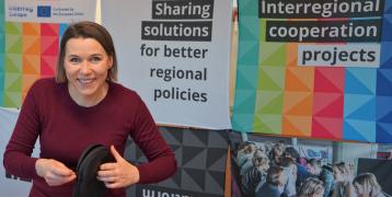 person smiling and standing in front of a colourful Interreg Europe stand and holding headphones