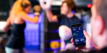 In front a man holding a phone showing data. In the backround two women are boxing.