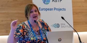 Portrait of ASTP CEO Laura McDonald Speaking at ASTP Annual Conference. In the background there is a banner with all the EU projects ASTP is part of. Laura has a laptop with a michrophone in front of her, and she is looking directly in the camera.