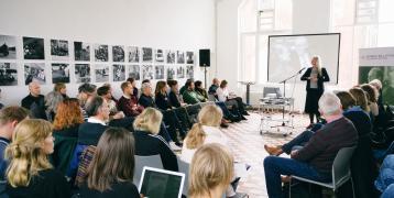 A group of people sitting in a room in an u-shape, a woman holds a speech